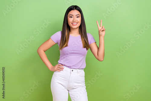 Photo of young optimistic korean lady wear purple t-shirt white pants show v-sign peace symbol clothes store isolated on green background
