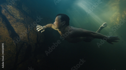 Young man free dives in the lake located in a forest and swims by underwater rocks