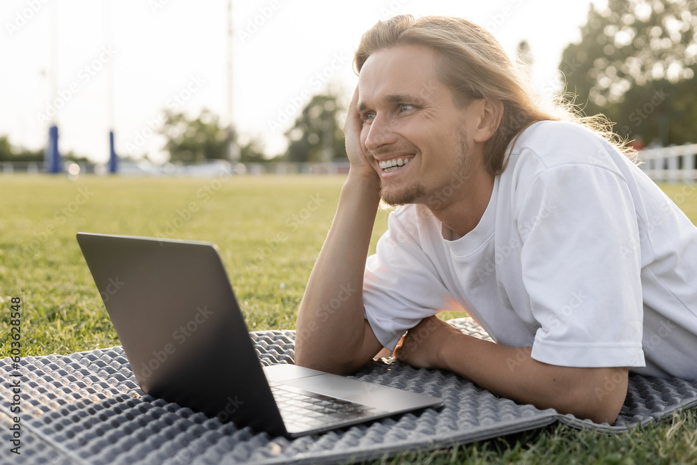 young cheerful yoga man looking away while lying near laptop on grassy stadium outdoors young cheerful yoga man looking away while lying near laptop on grassy stadium outdoors