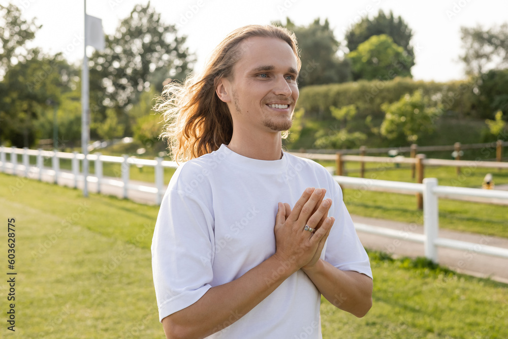 joyful long haired man in white t-shirt looking away while meditating with anjali mudra outdoors joyful long haired man in white t-shirt looking away while meditating with anjali mudra outdoors