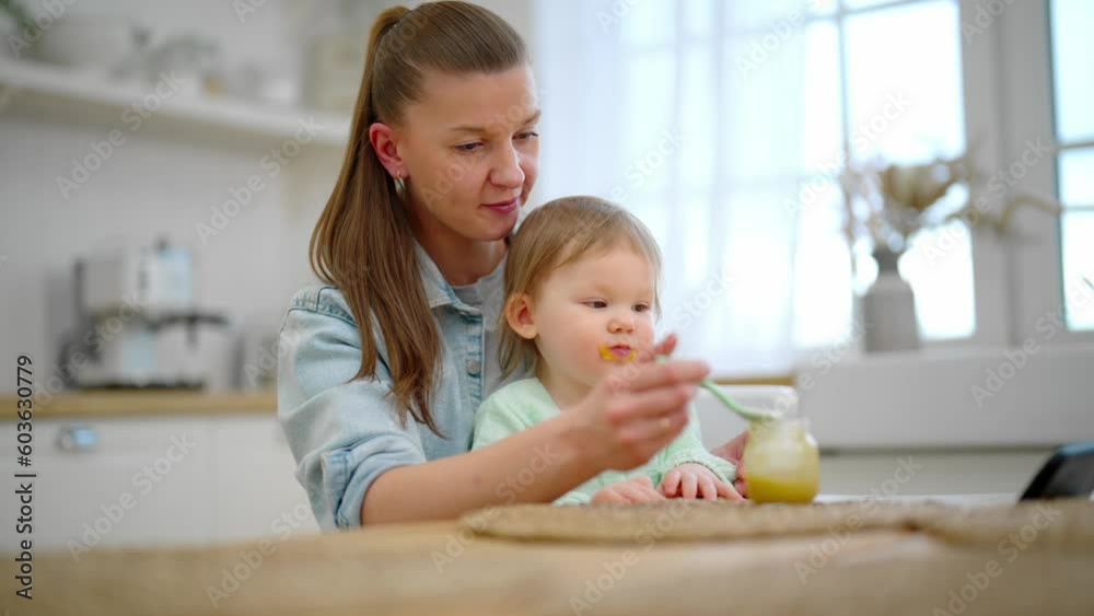 Mom trying to feed toddler baby vegetable puree from plastic spoon. Baby watching cartoons on smartphone sits on mother's knees at table on kitchen. Raising child, nutrition and motherhood concept. Mom trying to feed toddler baby vegetable puree from plastic spoon. Baby watching cartoons on smartphone sits on mother's knees at table on kitchen. Raising child, nutrition and motherhood concept.