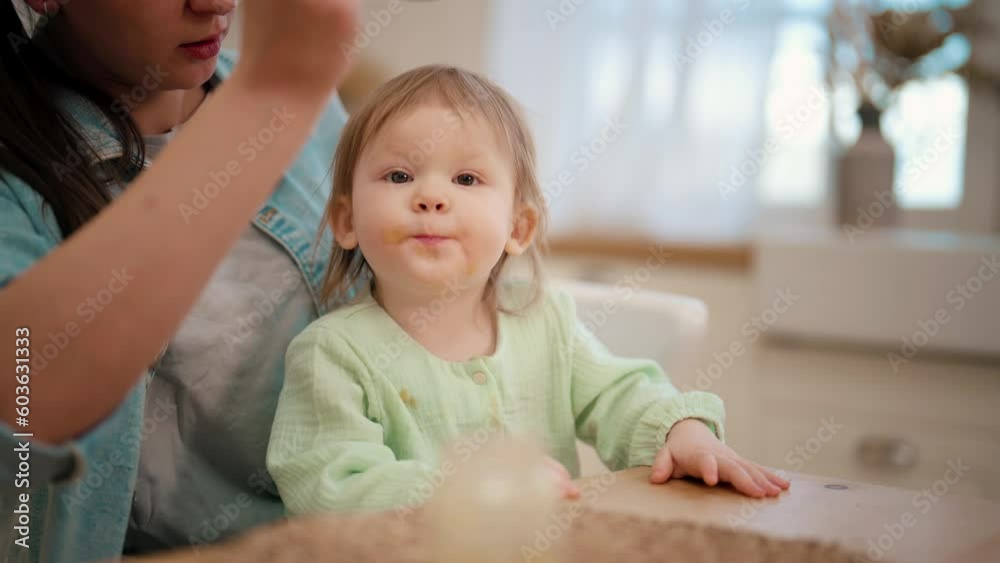 Mom feeds her baby playing with food. Toddler eats fruit or vegetable puree. Mother holds child on knees at table on kitchen. Raising child, kid's nutrition, motherhood, love care in family concept. Mom feeds her baby playing with food. Toddler eats fruit or vegetable puree. Mother holds child on knees at table on kitchen. Raising child, kid's nutrition, motherhood, love care in family concept.