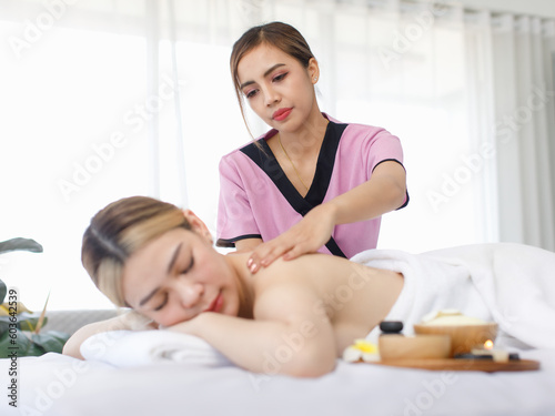 Portrait shot of Millennial Asian young beautiful relaxing resting nude naked woman laying lying down smiling look at camera on comfortable bed while female masseuse massaging shoulder and back