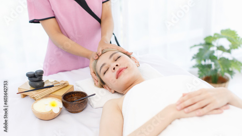 Millennial Asian young beautiful relaxing resting nude naked woman laying lying down on comfortable bed while unrecognizable female masseuse therapist preparing massaging head with oil and hot stones