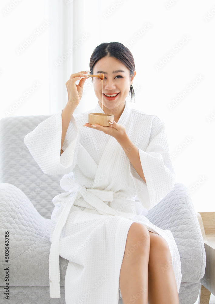 Millennial Asian beautiful pretty cheerful female model in white clean bathrobe sitting smiling on chair holding honey sugar wax bowl with stick ready to waxing skin by massaeuse in massage studio Millennial Asian beautiful pretty cheerful female model in white clean bathrobe sitting smiling on chair holding honey sugar wax bowl with stick ready to waxing skin by massaeuse in massage studio