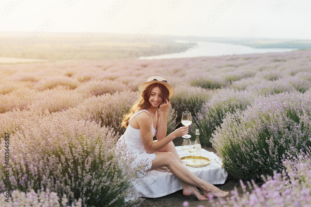 Young curly woman in white dress drinking wine in lavender field. Picnic on the field.