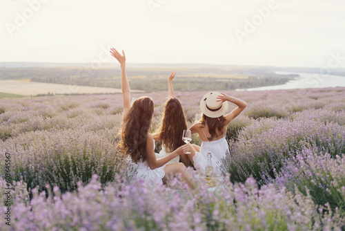 Obraz na płótnie Young female friends is having picnic with wine, cheese and fresh fruits on lavender field at sunset. w salonie