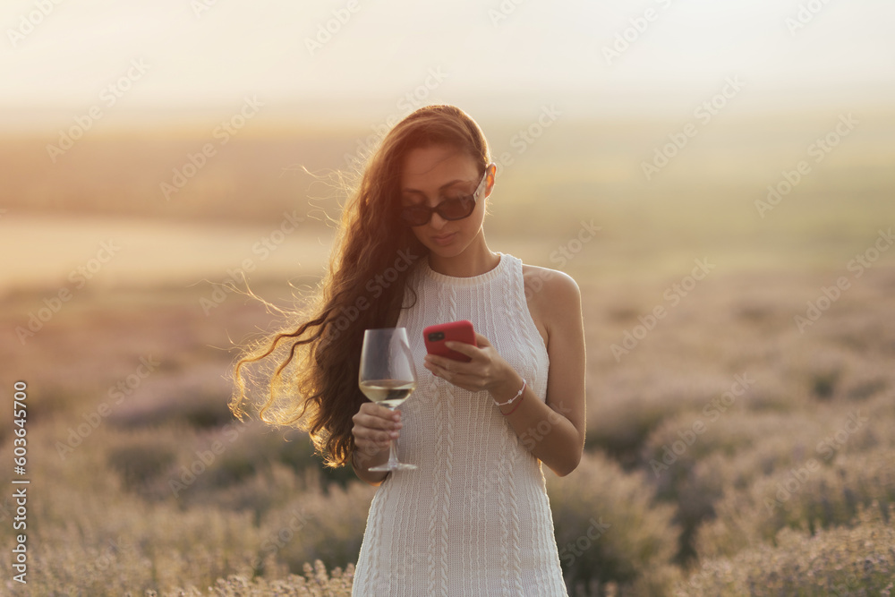 A woman in a white dress is using a smartphone and hold a glass of wine.