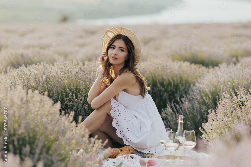 Beautiful woman in a straw hat and white dress sits on a picnic in a lavender field.