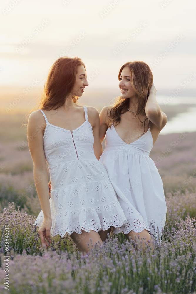 Two young happy sisters in white dresses stand in a field of lavender and looking each other.
