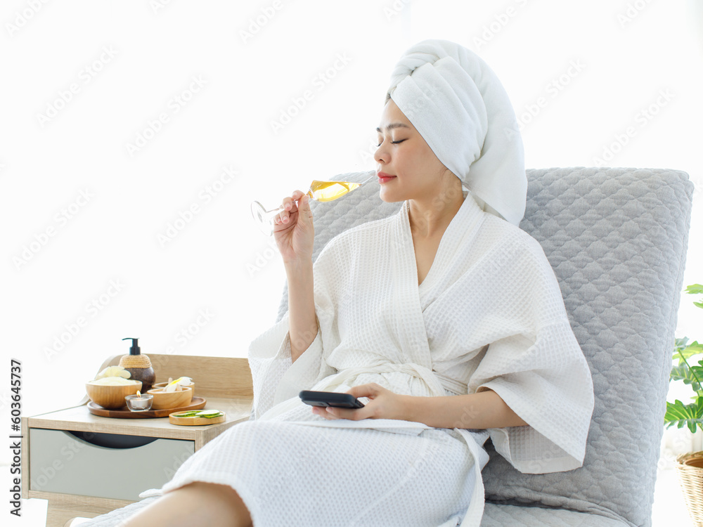 Millennial Asian female customer in white clean bathrobe and towel sitting on armchair drinking sparkling champagne while surfing internet via touchscreen smartphone waiting for masseuse to massaging Millennial Asian female customer in white clean bathrobe and towel sitting on armchair drinking sparkling champagne while surfing internet via touchscreen smartphone waiting for masseuse to massaging