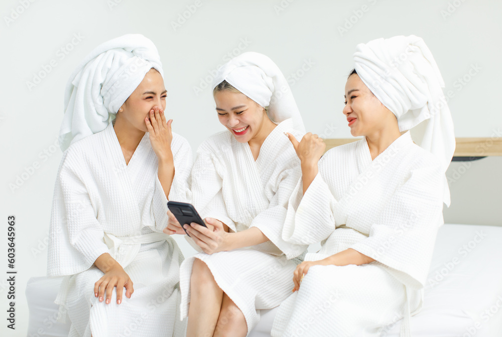 Millennial three Asian female customers friends in white clean bathrobes and towels have appointment at massage resort sitting on chair holding smartphone playing together after massaging