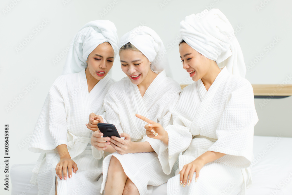 Millennial three Asian female customers friends in white clean bathrobes and towels have appointment at massage resort sitting on chair holding smartphone playing together after massaging Millennial three Asian female customers friends in white clean bathrobes and towels have appointment at massage resort sitting on chair holding smartphone playing together after massaging