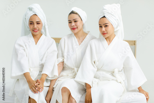Millennial three Asian female customers friends in white clean bathrobes and towels have appointment at massage resort sitting on chair chit chat talking having conversation together before massaging