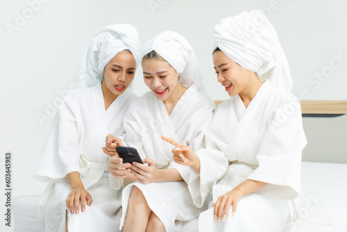 Millennial three Asian female customers friends in white clean bathrobes and towels have appointment at massage resort sitting on chair holding smartphone playing together after massaging