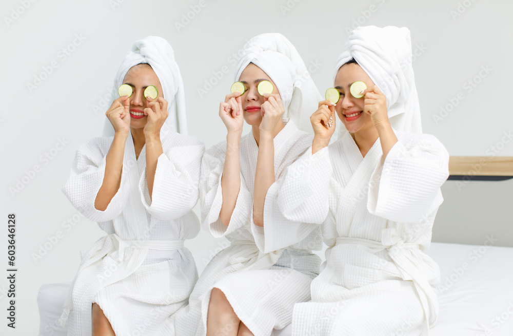 Millennial three Asian female customers friends in white clean bathrobes and towels have appointment at massage resort sit on chair holding sliced cucumbers placed on closed eyes treatment together Millennial three Asian female customers friends in white clean bathrobes and towels have appointment at massage resort sit on chair holding sliced cucumbers placed on closed eyes treatment together