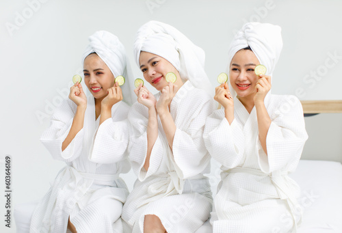 Millennial three Asian female customers friends in white clean bathrobes and towels have appointment at massage resort sit on chair holding sliced cucumbers placed on closed eyes treatment together