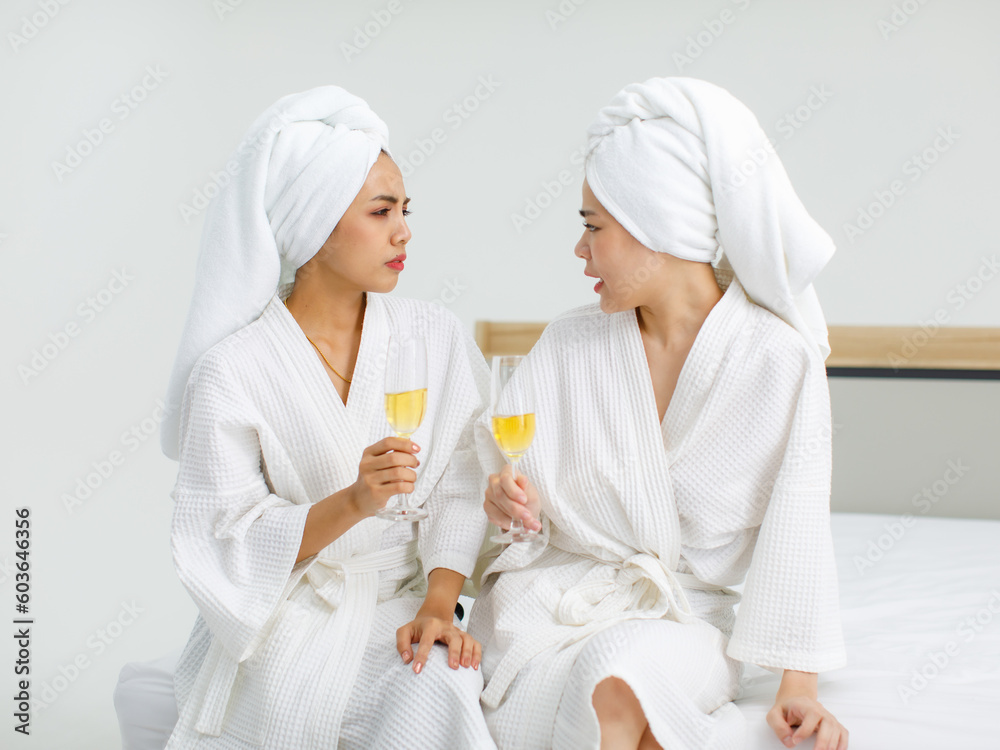 Millennial two Asian female customers friends in white clean bathrobes and towels have appointment at massage resort sitting on chair holding champagne glasses drinking together after massaging