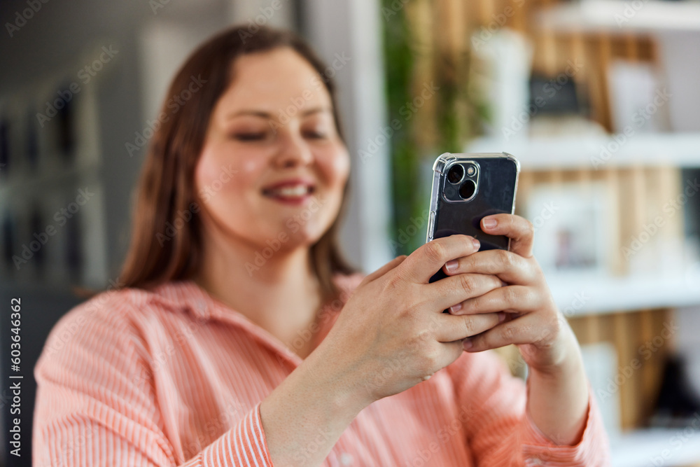 A focus on a smartphone, used by smiling plus-size woman while enjoying at home. A focus on a smartphone, used by smiling plus-size woman while enjoying at home.