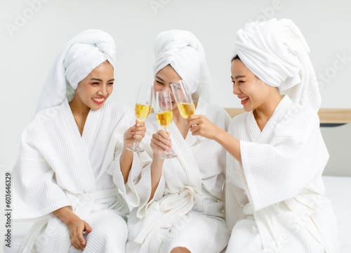 Millennial three Asian female customers friends in white clean bathrobes and towels have appointment at massage resort sitting on chair holding champagne glasses drinking together after massaging