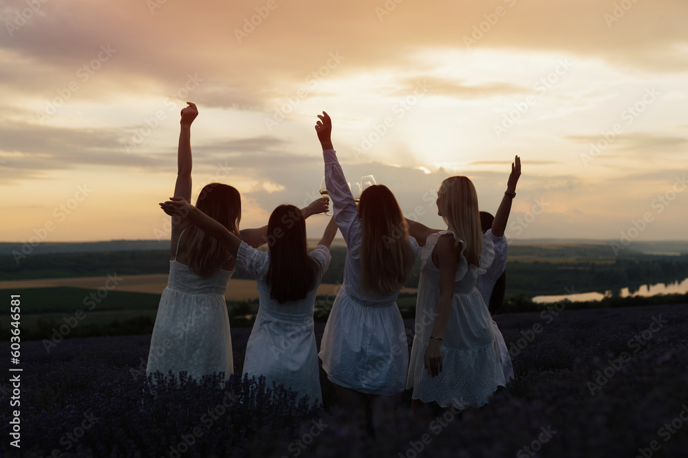 Silhouette of five women in white dresses while standing in a field with their arms in the air, with the sun setting behind them.