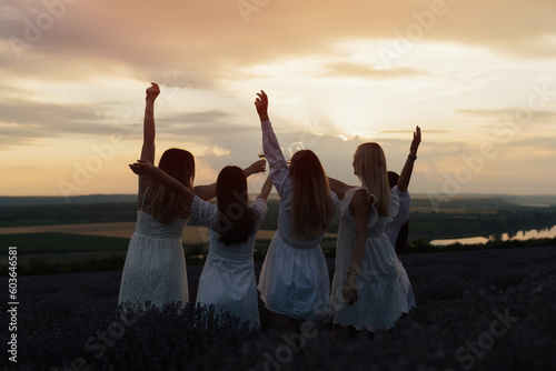 Silhouette of five women in white dresses while standing in a field with their arms in the air, with the sun setting behind them.