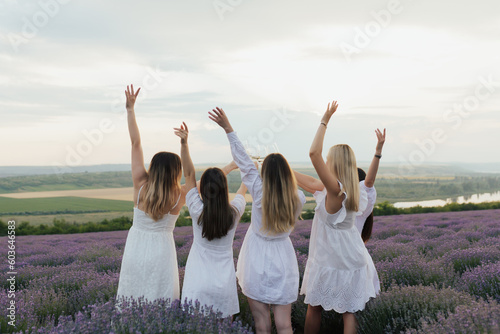 Back view of five women in white dresses are standing in a lavender field with their arms in the air.
