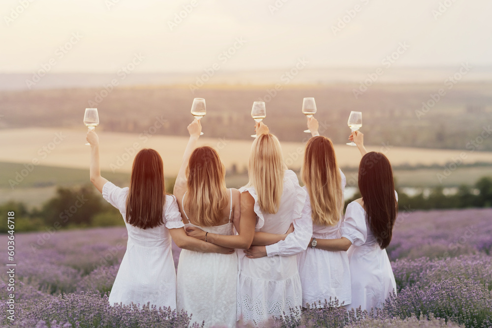 Back view of five women in white dresses hold up glasses of wine in a lavender field at sunset.