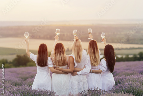 Obraz na płótnie Back view of five women in white dresses hold up glasses of wine in a lavender field at sunset. w salonie
