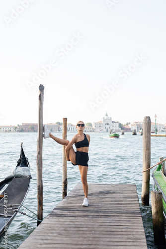 Flexible sportswoman in sunglasses, white sneakers, black crop top and shorts training on pier in Venice