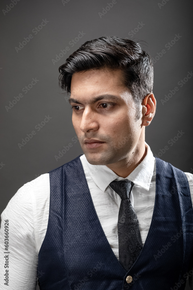 Studio shot of cheerful, young, handsome Indian male model in formal wear wearing white shirt, black tie, black coat against grey background. Male model. Fashion Portrait.