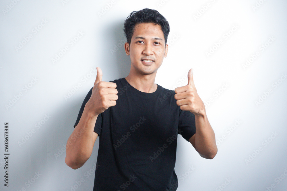 Young handsome Asian man wearing black tshirt over white background doing happy thumbs up gesture with hand. Approving expression looking at the camera showing success. Young handsome Asian man wearing black tshirt over white background doing happy thumbs up gesture with hand. Approving expression looking at the camera showing success.