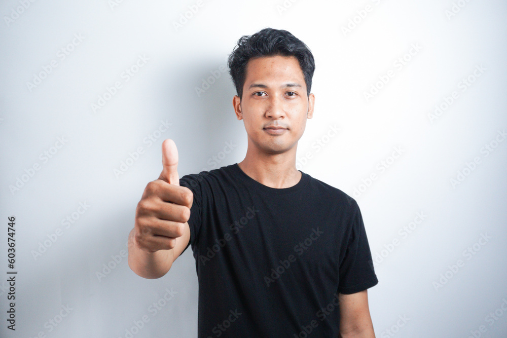 Young handsome Asian man wearing black tshirt over white background doing happy thumbs up gesture with hand. Approving expression looking at the camera showing success. Young handsome Asian man wearing black tshirt over white background doing happy thumbs up gesture with hand. Approving expression looking at the camera showing success.