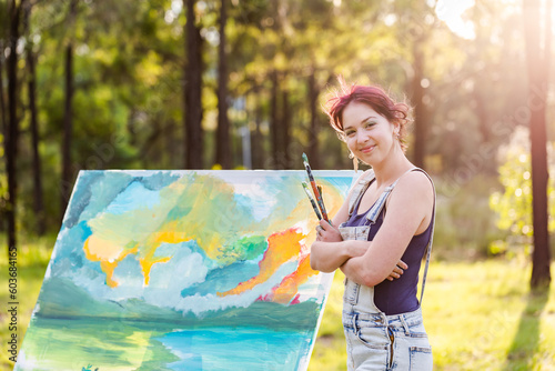 Smiling portrait of happy young woman artist in forest clearing with beginning of painting