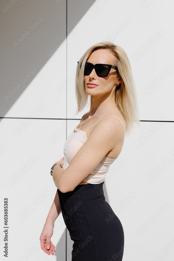 Adorable blonde adult woman, wearing in top and pants, standing at white wall looking at camera