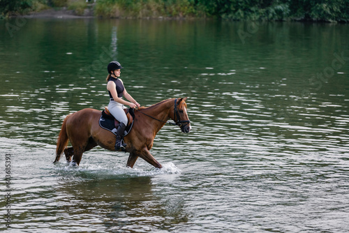 Scenic view of a female horseback rider on a beautiful red chestnut horse riding in the river towards the setting sun. Animals, people, and environment concept.