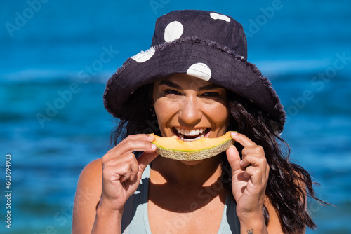 Close up of Young woman wearing full brim hat and eating rockmelon on the beach on clear sunny day photo