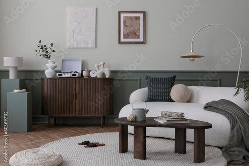 Warm and cozy living room interior with mock up poster frame, boucle sofa, wooden sideboard, coffee table, stylish lamp, gramophone, vase with leaves and personal accessories. Home decor. Template.