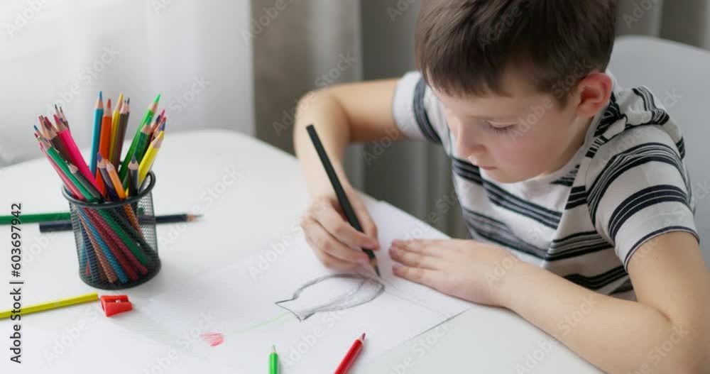 A child boy draws on white paper with colored pencils while sitting at a table A child boy draws on white paper with colored pencils while sitting at a table