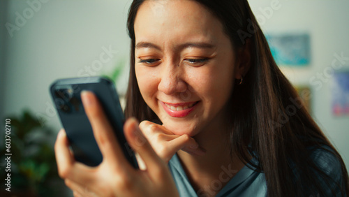 Portrait of Happy beautiful Asian woman sit on sofa hand holding mobile phone chatting and playing social media. Young female looking at smartphone cellphone browsing internet on sofa home living room