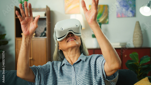 Old beautiful Asian elderly woman putting on Virtual Reality metaverse headset technology exploring playing VR world game amazed, surprised and fun emotional sit on sofa at home in cozy living room.