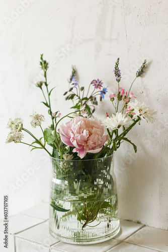 Stylish bouquet with peony and wildflowers on tile shelf on rustic wall background in modern room. Beautiful flowers in glass vase gathered from garden, summer floral arrangement in home.