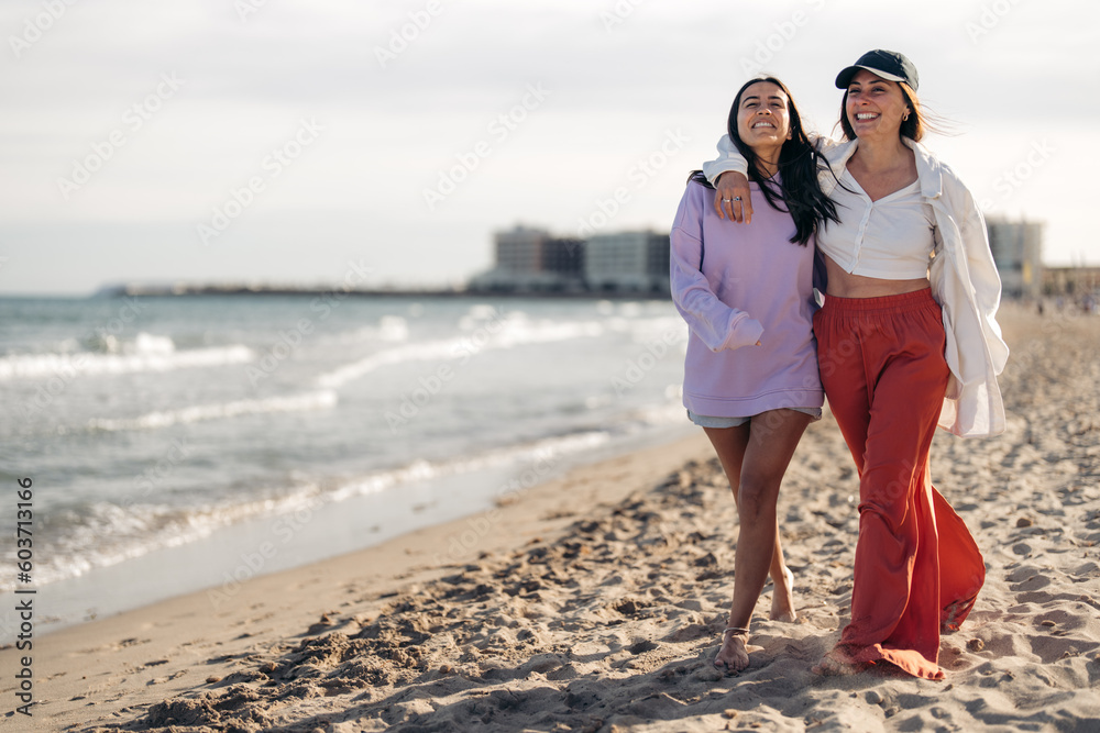 Two happy smiling girlfriends walking by the ocean on sandy beach with arms around each other. Cheerful young women in casual clothes barefoot spending time on the beach. Two happy smiling girlfriends walking by the ocean on sandy beach with arms around each other. Cheerful young women in casual clothes barefoot spending time on the beach.