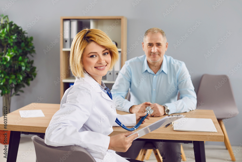 Obraz premium Portrait happy young female doctor during medical consultation at work. Pretty blond woman general practitioner in white coat sitting at desk with patient in background, looking at camera, and smiling