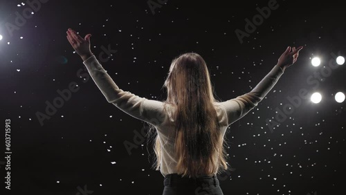 Back view of caucasian young woman keeping hands up from happiness after successful inspirational seminar. Multiple spotlights light black stage with professional business mentor.