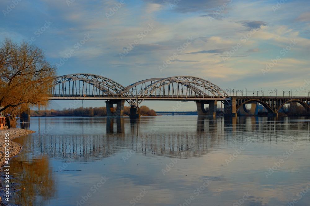 Scenic spring landscape Dnipro River in Kyiv during sunset. Bridge reflected in tranquil water of Dnieper River. Blue sky with white clouds in the background. Nature concept