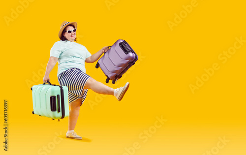 Happy funny woman going on summer vacation. Cheerful fat overweight woman in T shirt, shorts, sun hat and sunglasses holding green and purple travel suitcases and walking isolated on yellow background