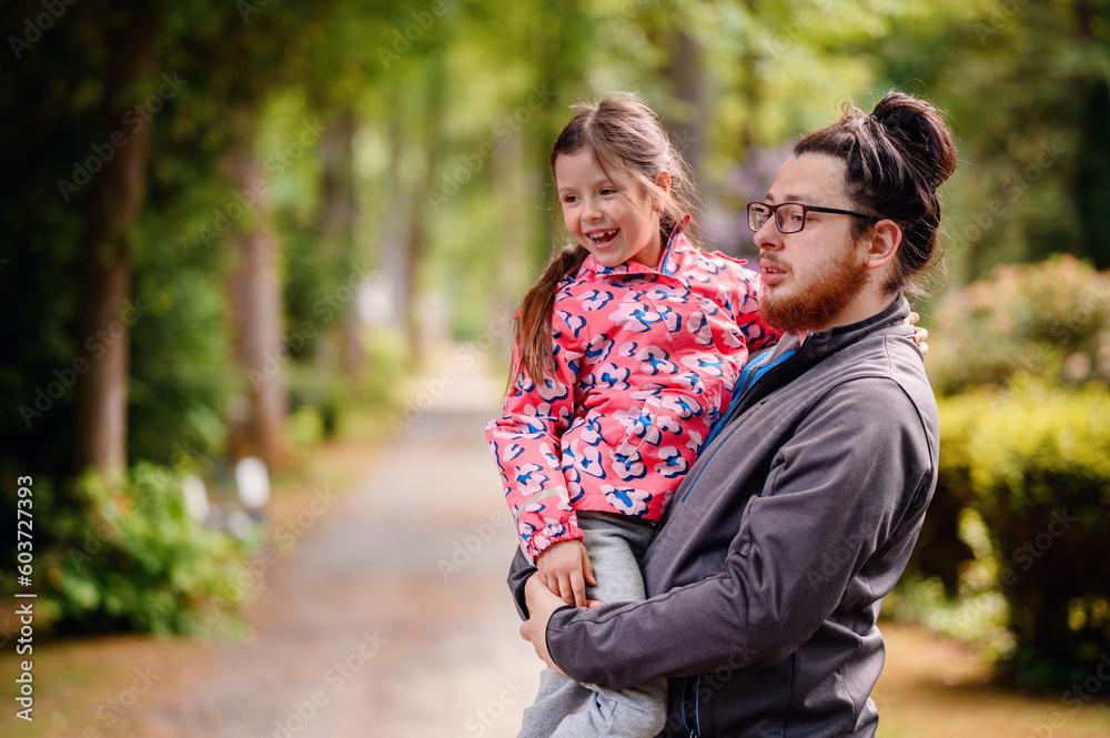 Fototapeta premium Young long hair bearded man in glasses standing with junior girl holding on hands on road in park