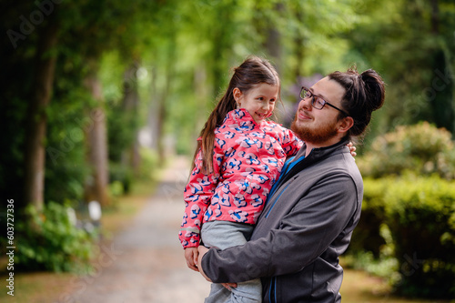 Young long hair bearded man in glasses standing with junior girl holding on hands on road in park