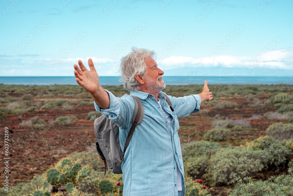 Happy active senior man in outdoors hiking day holding backpack looking at the sky with open arms enjoying freedom